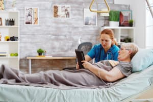 Female caregiver helps an old disabled woman lying in hospital bed to use a digital tablet PC